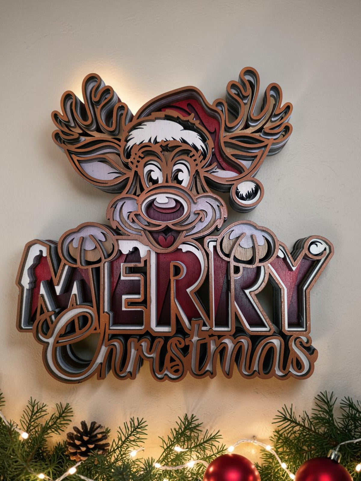 Layered wooden Merry Christmas reindeer wall art styled with evergreen branches, ornaments, and warm holiday lights